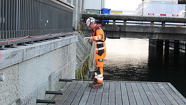 Sicherheitsbeauftragter knotet Seile an Ankerpunkte. Steht auf Holzsteg und im Hintergrund ist das Wasser der Limmat zu sehen.
