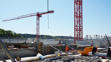 Baustelle mit mehreren Bauarbeiter:innen im Einsatz auf einer Betonbodenplatte, umgeben von Schalungselementen, Baumaterial und Geräten. Zwei grosse rote Turmdrehkrane ragen in den blauen Himmel, im Hintergrund eine grüne Baumreihe und ein Gerüst entlang der Betonmauer.