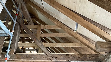 Blick in einen Dachstuhl mit Holzbalken und Verkleidung, links ist ein Gerüst zu sehen. Eine Kombination aus altem vorhandenem Holz mit neuem Holzelementen. 