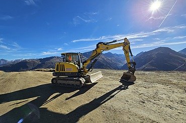 Gelber STRABAG Bagger auf einer geraden Sandfläche. Im Hintergrund blauer Himmel mit strahlendem Sonnenschein und einem Bergpanorama