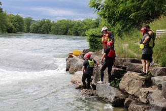 Teilnehmende eines Wasserrettungskurses stehen in Schutzkleidung auf Felsen am Flussufer und beobachten die Strömung des Wassers.