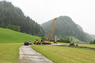 Baustelle im Grünen mit gelbem Kran, der einen Filterbrunnen für den Einbau aufstellt, und einem Bagger an einem Graben.