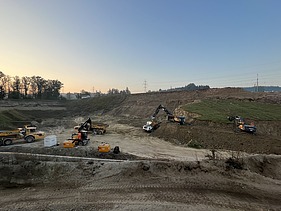 Mehrere Baumaschinen und Lastwagen arbeiten auf einer grossen Erdbaustelle im Burgaurfeld, im Hintergrund sind Hänge und Bäume zu sehen.