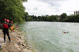 Teilnehmende eines Wasserrettungskurses stehen in Schutzkleidung am Ufer eines Flusses, während eine Person im Wasser eine Rettungsübung in der Strömung durchführt.