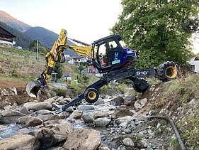 Arbeiten im Bach - STRABAG Ingenieurbau Ein STRAAG Schreitbagger im Bachbett des Aclettbachs bei Disentis während eines Wasserbauprojekts. Im Hintergrund ein begrünter Baum