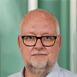 Technischer Gruppenleiter im Anzug lächelt in die Kamera. Im Hintergrund ist ein Lift zu sehen.