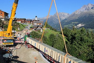 Ein großer Pneukran hebt einen langen Holzträger langsam in die Höhe; darunter stehen Arbeiter und im Hintergrund sieht man ein Bergdorf vor einer beeindruckenden Alpenkulisse.