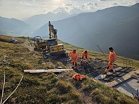 3 STRABAG-Bauarbeiter führen Handarbeiten an einem neuangelegten Weg aus. Im Hintergrund öffnet ein Bagger den Boden für den zukünftigen Weg und ein Tumber transportiert den Aushub ab. Im Hintergrund sieht man die Sonne durch eine Wolke durchstrahlen und eine Berglandschaft. 