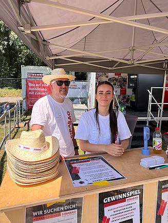 Zwei STRABAG-Lernende in weissen STRABAG T-Shirts am STRABAG Stand unter einem Bauzelt.