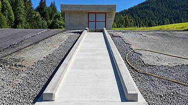 Perspektive entlang frisch betonierter Schussrinne aufwärts mit Betriebsgebäude im Hintergrund