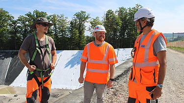 Drei Männer in Schutzkleidung stehen auf der Baustelle beisammen und sprechen miteinander.