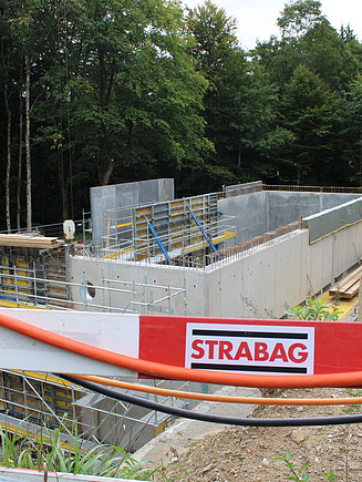 Baulatten mit STRABAG-Logo vor der Baugrube, im Hintergrund der Rohbau des neuen Wasserreservoirs Kopfholz in Rüschlikon