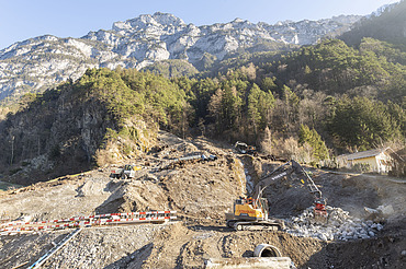 Ein Bagger der STRABAG räumt Geröll an einem steilen Hang unterhalb einer Felswand weg. Baustelle zur Hangsicherung im Gebirge.