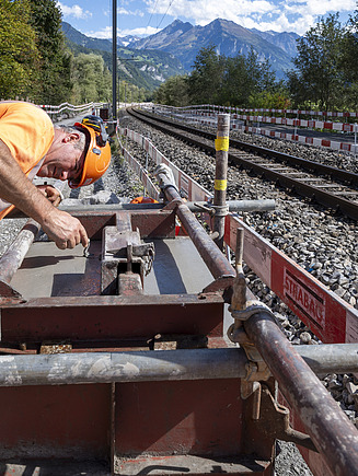 Ein Bauarbeiter in oranger Schutzkleidung arbeitet zwischen den Bahngleisen. Rundherum verlaufen provisorische Leitungen und Absperrgitter, im Hintergrund sieht man die langen Gleisanlagen