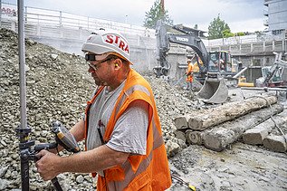 Der Polier in der Baugrube beim Vermessen am Lasermessgerät im Hintergrund sieht man das restliche Baustellenpersonal und zwei Bagger