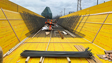  Ein Kran der STRABAG hebt nachts ein langes Brückenteil über Bahngleise. Die Baustelle ist hell erleuchtet.