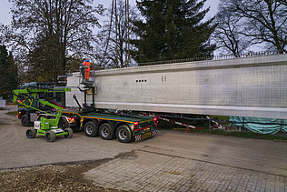 Ein grosser Turnhallenträger liegt auf einem langen Transportfahrzeug auf einer Strasse.