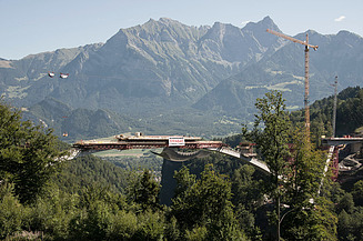Blick auf eine im Bau befindliche Brücke von STRABAG in alpiner Landschaft mit Kränen und Baugerüst.