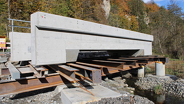 Ein grosses Betonfertigelement liegt auf provisorischen Stahlträgern über einem ausgehobenen Graben. Im Hintergrund stehen Bäume.