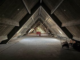 Historischer Dachstuhl mit markanter, dreieckiger Holzkonstruktion in A-Form. In der Mitte des Raumes steht ein roter Baustellenwagen, rechts eine Leiter und Baumaterial. Durch seitliche Fensteröffnungen fällt etwas Tageslicht ins Innere. Der Raum befindet sich noch im Umbau.