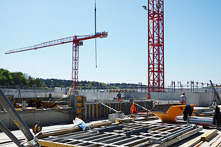 Baustelle mit mehreren Bauarbeiter:innen im Einsatz auf einer Betonbodenplatte, umgeben von Schalungselementen, Baumaterial und Geräten. Zwei grosse rote Turmdrehkrane ragen in den blauen Himmel, im Hintergrund eine grüne Baumreihe und ein Gerüst entlang der Betonmauer.