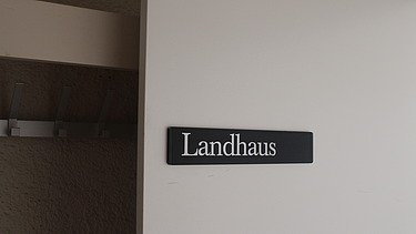 Nahaufnahme vom Türschild mit der Aufschrift «Landhaus» in weisser Schrift auf schwarzem Hintergrund, montiert an einer hellen Wand. Links im Bild ist ein Teil einer Garderobe mit Haken zu sehen.
