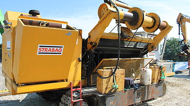 Gelbe Baumaschine mit mehreren Walzen und Anbaugeräten steht auf einer Baustelle im Hintergrund sind weitere Maschinen und Material zu sehen.