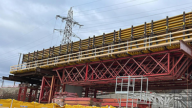  Ein Kran der STRABAG hebt nachts ein langes Brückenteil über Bahngleise. Die Baustelle ist hell erleuchtet.