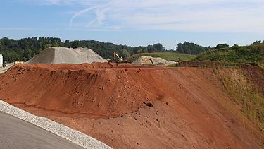 Erddeponie mit grossen Aushubmengen im Vordergrund eine rötliche Böschung, im Hintergrund sind Maschinen und Ablagerungen zu sehen.