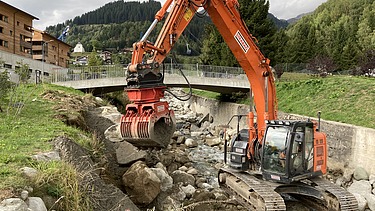 Arbeiten im Bach - STRABAG Ingenieurbau Bagger der STRABAG arbeitet an einer Flussverbauung am Aclettbach bei Disentis. Eine Brücke führt über den Bach.