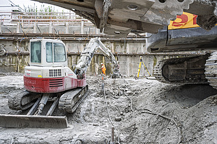 Bild aus der Baugrube im Vordergrund ein Minibagger im Hintergrund ein grosser Bagger bei dem die Schaufel ins Bild rein ragt 