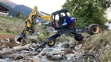 Arbeiten im Bach - STRABAG Ingenieurbau Ein STRAAG Schreitbagger im Bachbett des Aclettbachs bei Disentis während eines Wasserbauprojekts. Im Hintergrund ein begrünter Baum