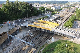 Fertig eingesetzte Bahnbrücke in Küssnacht. Die Brücke führt über mehrere Bahngleise. Im Hintergrund sind Strassen, Gebäude und ein Bahnhof zu erkennen.