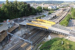 Fertig eingesetzte Bahnbrücke in Küssnacht. Die Brücke führt über mehrere Bahngleise. Im Hintergrund sind Strassen, Gebäude und ein Bahnhof zu erkennen.