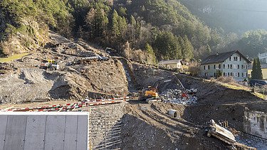 STRABAG-Baustelle am Hang: Volvo-Bagger und Dumper bei Erdarbeiten neben einer grossen Betonmauer in bergiger Umgebung.