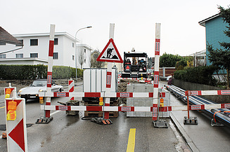 Baustelle auf einer Wohnstrasse mit rot-weissen Absperrungen. Ein STRABAG-Fahrzeug steht zentral im Bild, dahinter ein Bagger. Entlang des Gehwegs verlaufen Werkleitungen, links parkt ein Auto.