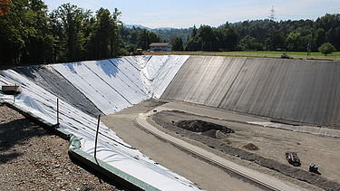 Blick auf eine grosse, asphaltierte Steilböschung mit hellem und dunklem Belag unten verläuft ein Baustellenweg, im Hintergrund Wald und Landschaft.