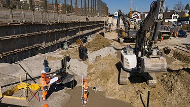 Rüschlikon Bahnhof Süd – STRABAG Tiefbau und Spezialtiefbau Drohnenaufnahme der Baugrube von vorne, zu sehen sind mehrere Bagger, im Vordergrund Bauarbeiter bei Vermessungen