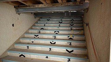 Denkmalgeschützte Treppe, deren Stufen provisorisch mit Schutzfolie abgedeckt sind. Darüber verlaufen offene Deckenbalken aus Holz sowie Stahlträger. Rechts an der Wand verläuft ein sichtbares Elektrokabel.