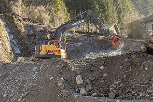 Gelber STRABAG-Bagger von Volvo sortiert mit einem Greifer grosse Steine auf einer Hangbaustelle im Gebirge.