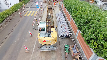 Luftaufnahme einer Baustelle mit offener Baugrube und einem Bagger. Die Baugrube ist mit Absperrungen und Verkehrsschildern gesichert, während Werkleitungen und Baumaterialien entlang der Strasse gestapelt sind. Im Hintergrund sind Bauarbeiter bei der Installation und Verlegung von Leitungen tätig. Die Baustelle befindet sich an einer Strasse mit markierten Fussgängerüberwegen und einer geregelten Verkehrsführung. 