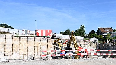Mehrere Baufahrzeuge in einer Baugrube bei der Arbeit. Im Hintergrund ein weisser STRABAG Container mit der Aufschrift STR in grossen roten Buchstaben.