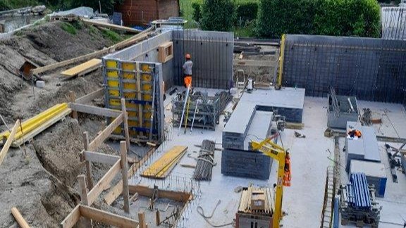 Eine Baugrube mit den Fundamenten und ersten Mauern eines 3-Familienhauses, ein Bauarbeiter bei der Arbeit ist ebenfalls zu sehen. 