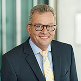 Kaufmännischer Bereichsleiter im Anzug lächelt in die Kamera. Im Hintergrund ist ein Lift zu sehen.