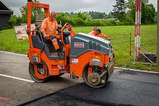 Ein Arbeiter fährt eine orange Strassenwalze und verdichtet frischen Asphalt auf einer Strasse.