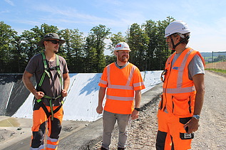 Drei Männer in Schutzkleidung stehen auf der Baustelle beisammen und sprechen miteinander.