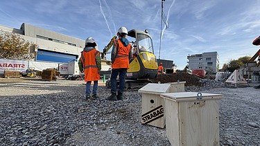 Zwei Schüler:innen warten vor dem Mini-Bagger in Baustellenweste und Baustellenhelme bis sie selbst ans Steuer dürfen