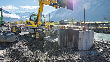 Ein orangener Bagger arbeitet auf einer Baustelle vor einer alpinen Bergkulisse. Neben dem Bagger steht ein grosser, runder Betonfundamentkörper, umgeben von Schotter und Erde. 