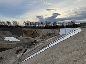 Blick über ein grosses Erdgefälle mit Baumaschinen unter einem wolkigen Himmel im Hintergrund sind Bäume zu sehen.