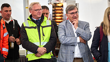 Mehrere Personen stehen bei einer Veranstaltung zusammen. Eine Person in einer gelben Sicherheitsjacke und eine andere im grauen Sakko stehen im Vordergrund und hören aufmerksam zu.